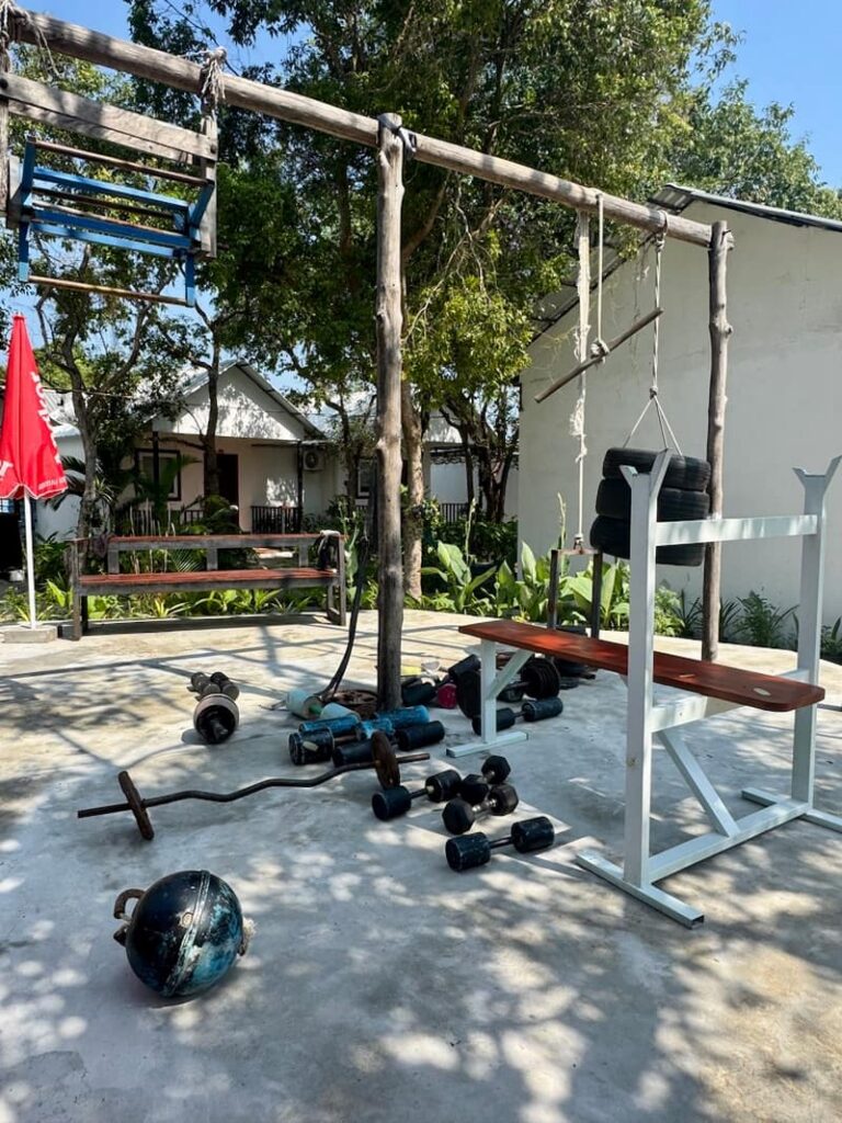 Open-air outdoor gym setup with homemade weights, a workout bench, and pull-up bars in a tropical resort setting.