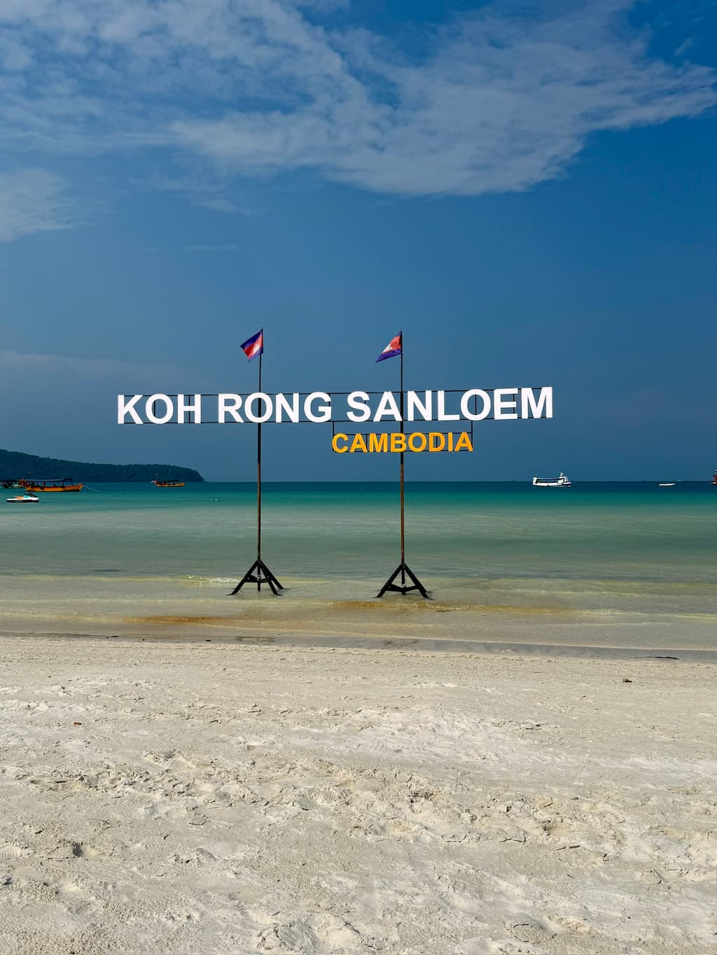 Large "KOH RONG SANLOEM CAMBODIA" sign standing on a white sand beach overlooking the turquoise sea under a blue sky.