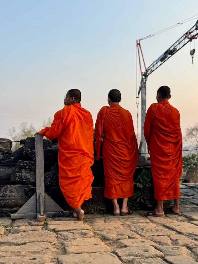 Three Buddhist monks in orange robes stand looking out from a stone terrace near a construction crane.