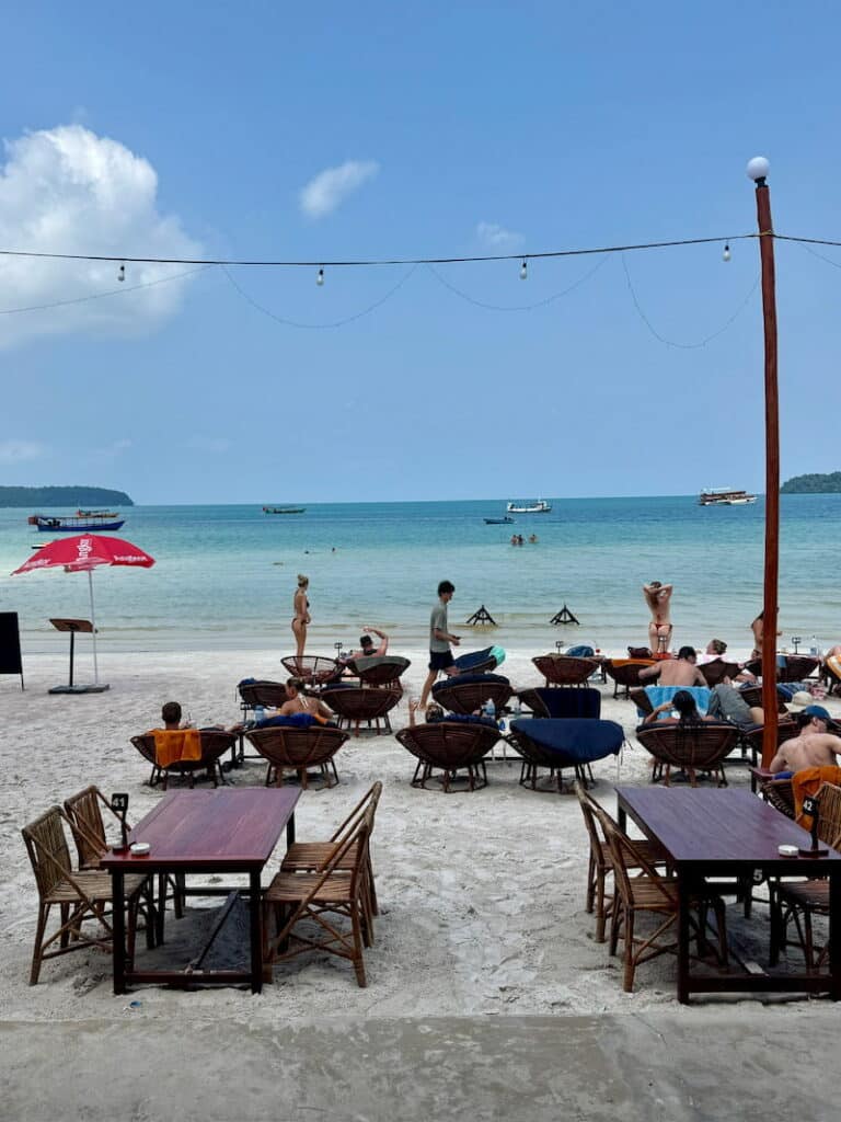 Open-air beach restaurant setup with wooden tables and chairs on the sand overlooking the turquoise sea and distant boats.