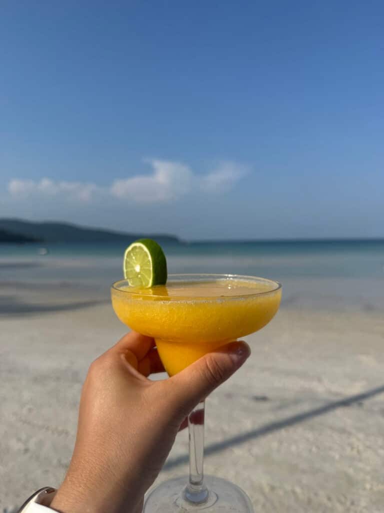 Hand holding a refreshing tropical cocktail garnished with a lime slice against a backdrop of a white sand beach and turquoise ocean.