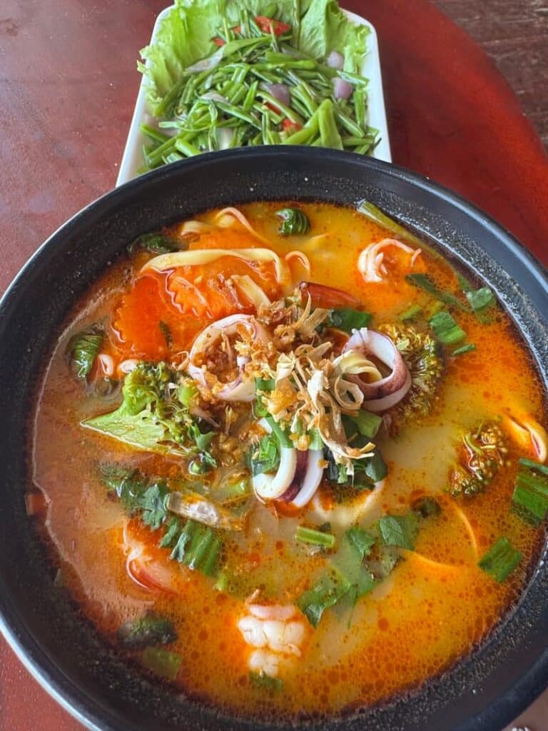 Spicy seafood noodle soup with squid, shrimp, and herbs served alongside a side of stir-fried morning glory.