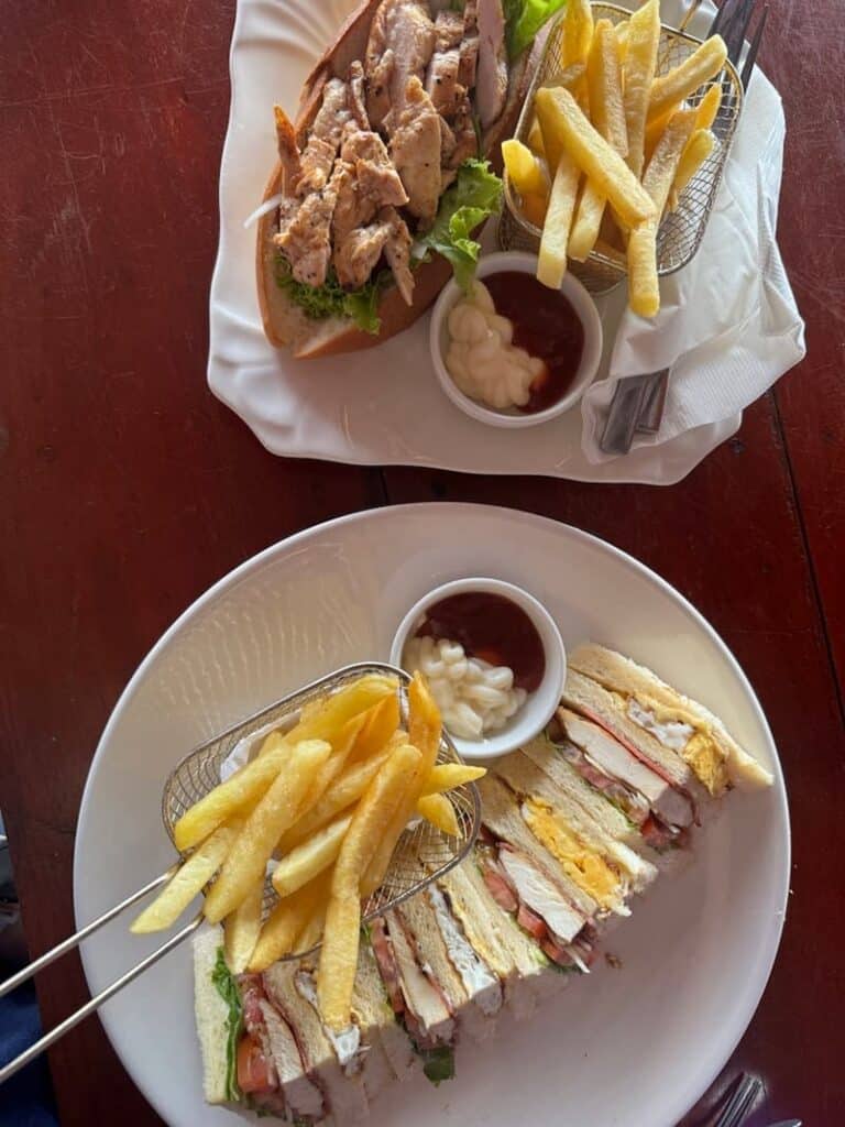 A grilled chicken baguette sandwich and a sliced club sandwich served with golden french fries and dipping sauces.