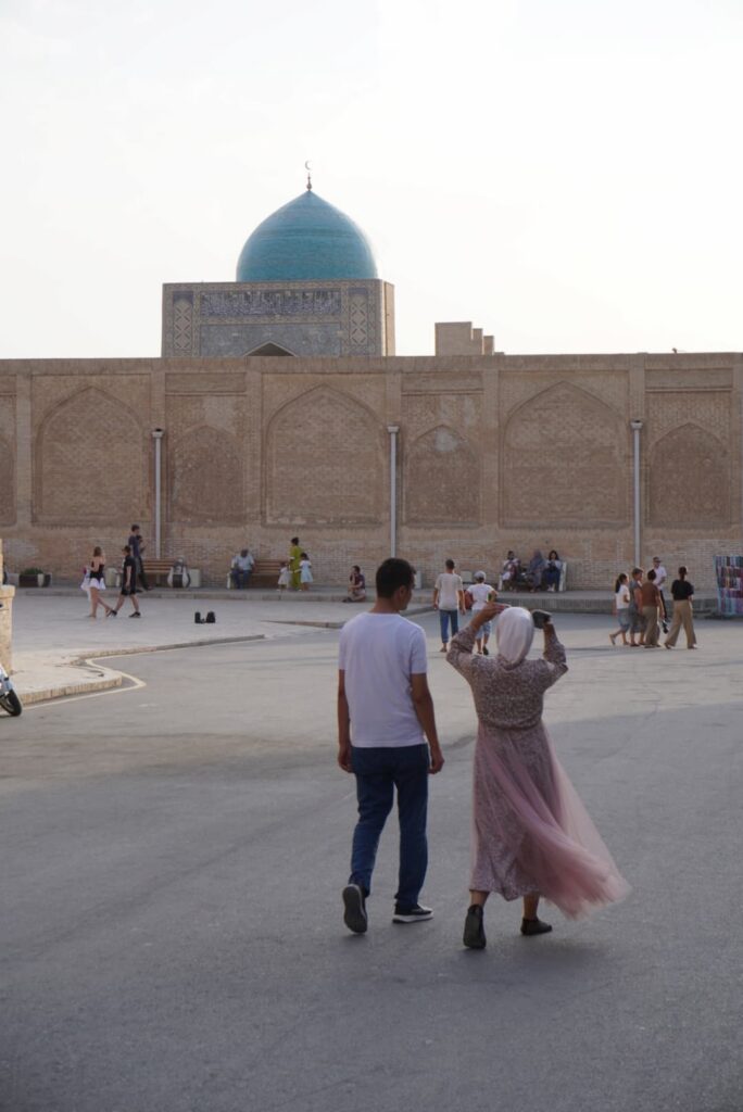 A local couple walking toward the historic Poi Kalyan complex in Bukhara, with the woman wearing a vibrant pink dress.