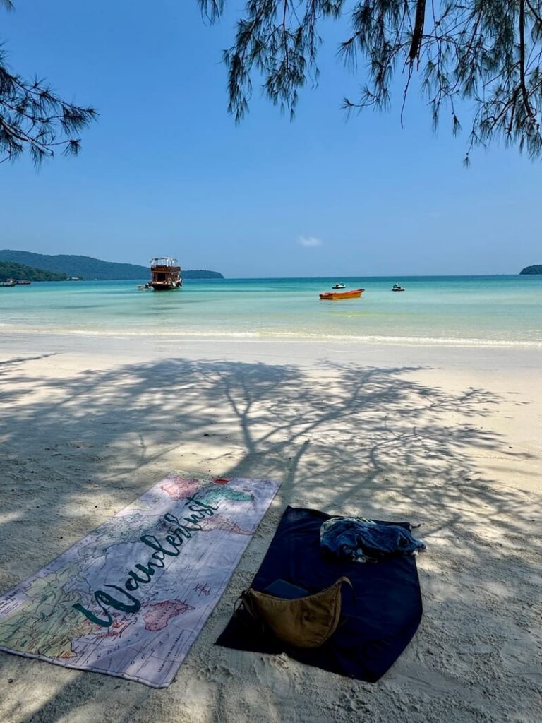 A "Wanderlust" beach towel and a dark mat spread out on the white sand of Saracen Bay under tree shade, overlooking the turquoise ocean and boats.