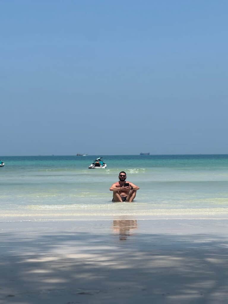 Ale sitting cross-legged in the shallow turquoise water at Saracen Bay, looking at his phone with a jet ski in the distance, as featured in our guide about how to spend 2 days in koh rong samloem.
