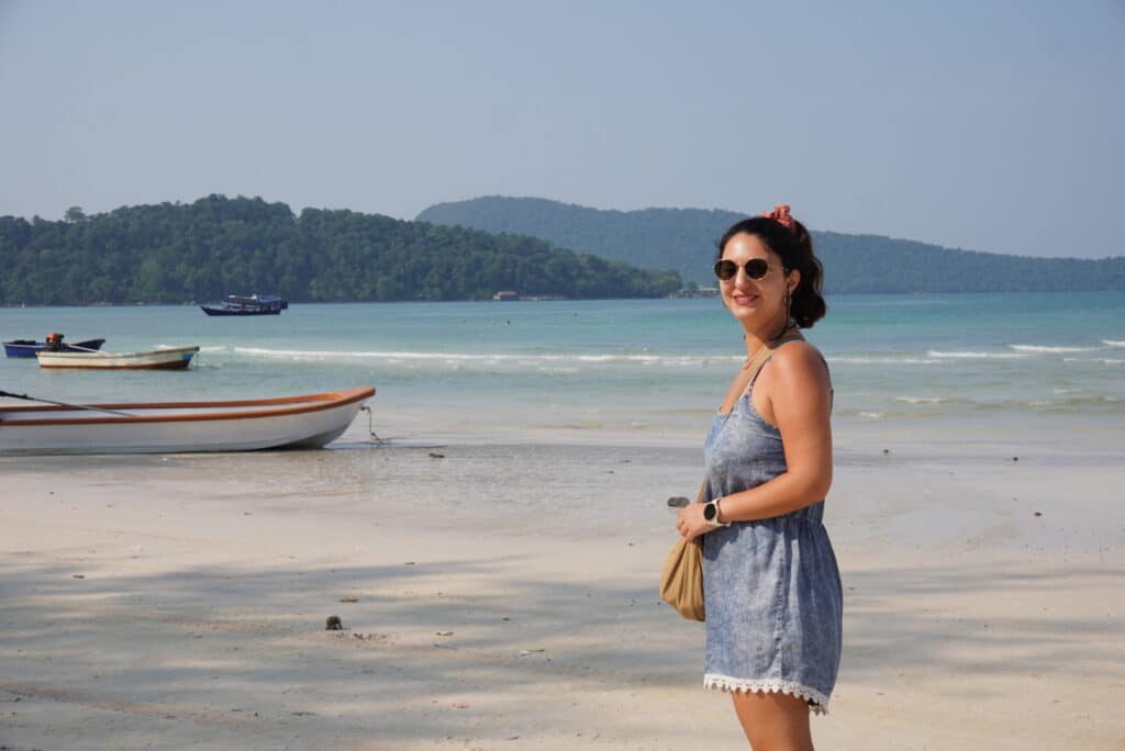 Eleni standing on the white sand beach at Saracen Bay wearing sunglasses, with turquoise water, small boats, and green hills in the background.