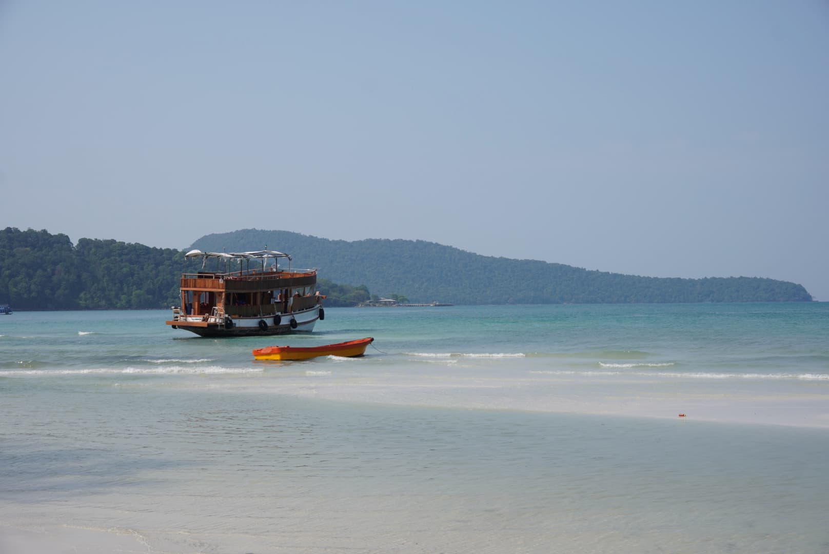 Wooden tour boat and small orange canoe floating in the crystal-clear turquoise water of Saracen Bay, with white sand and lush green hills in the background.