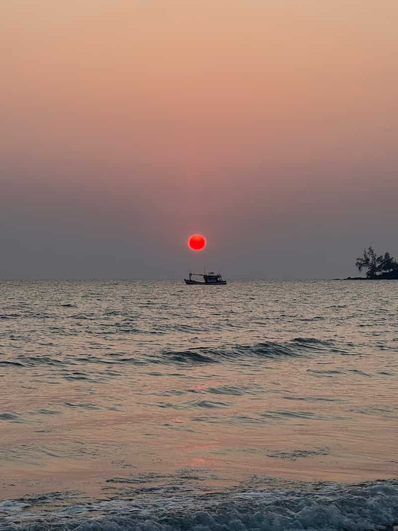 A vibrant red sun setting over the calm ocean with a small boat on the horizon and gentle waves in the foreground.