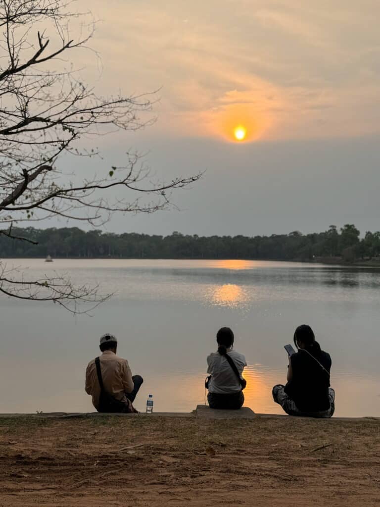 Three people sit on the edge of a lake at sunset, watching the golden sun reflect on the water within the Angkor complex.