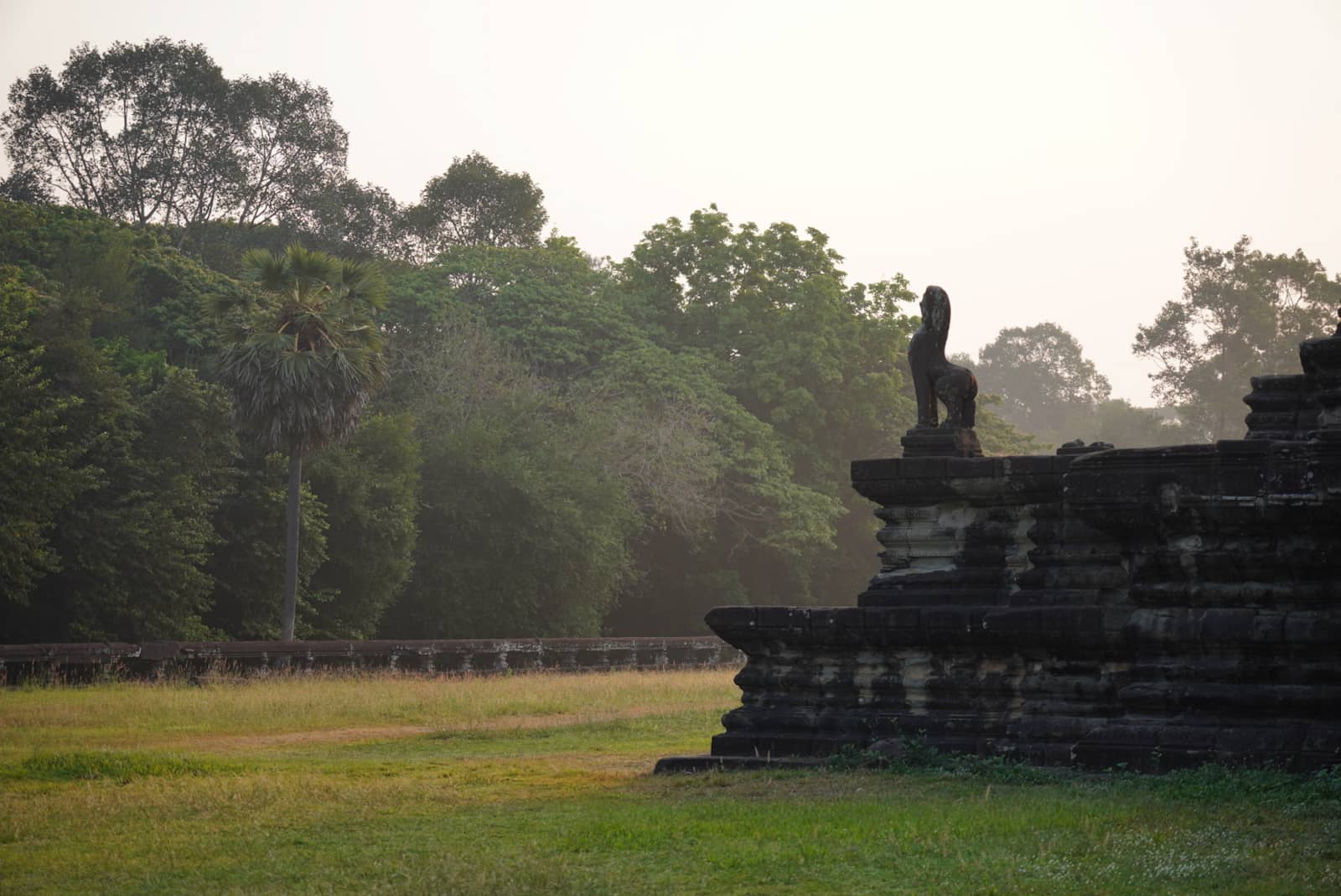 An ancient stone guardian statue perched on a weathered temple structure overlooking a grassy lawn and lush green trees.