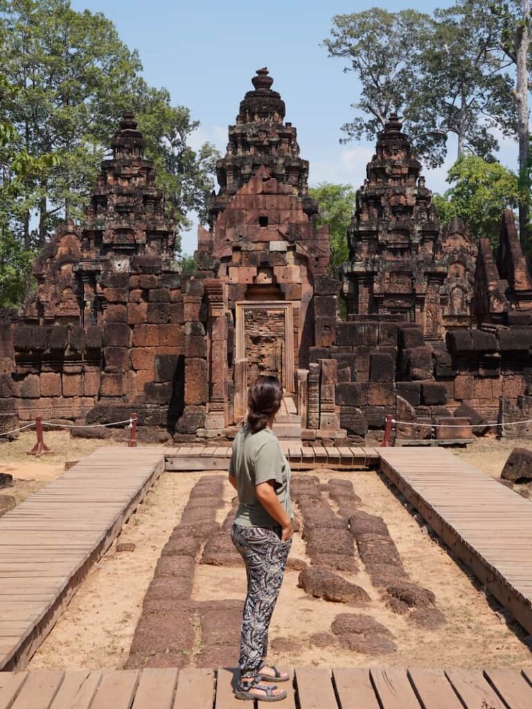 A visitor standing on a wooden walkway admiring the intricate red sandstone carvings of the Banteay Srei temple in Siem Reap.