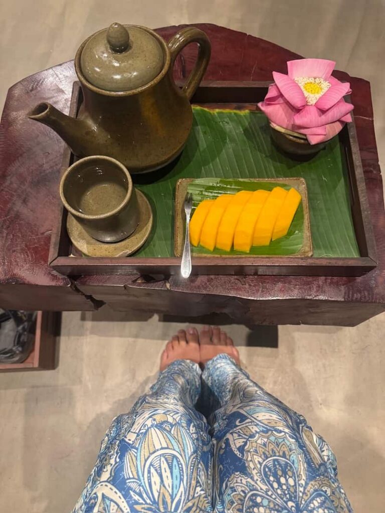 A relaxing tray with a ceramic teapot, a cup of tea, sliced fresh mango on a banana leaf, and a folded pink lotus flower at Bodia Massage in Phnom Penh.