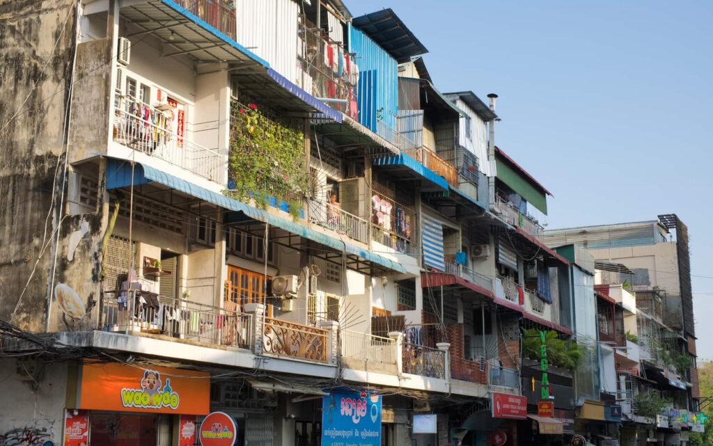 A row of multi-story urban residential and commercial buildings with balconies, hanging laundry, and storefronts along a street in Phnom Penh.