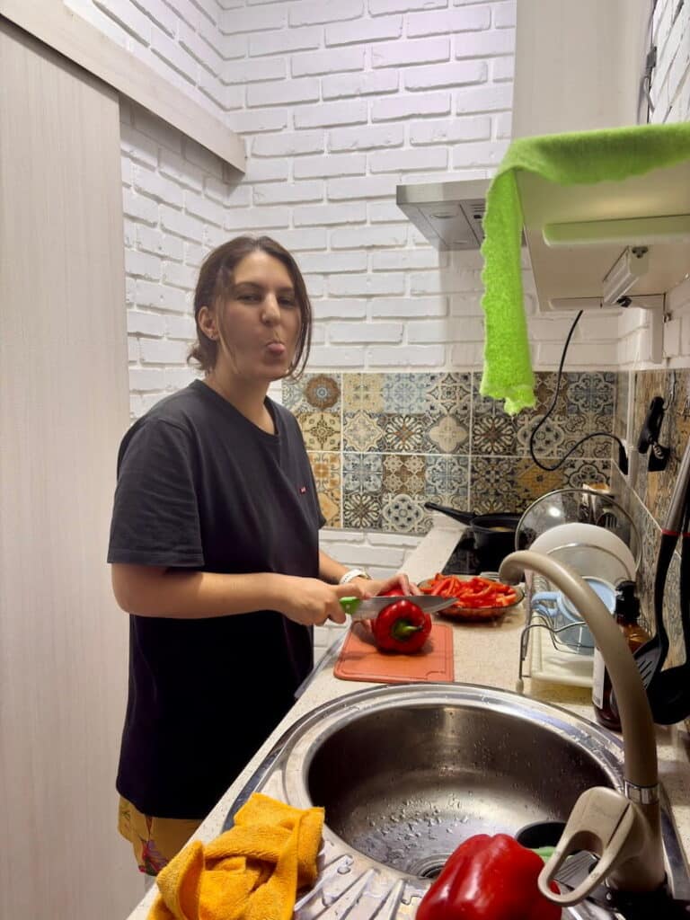 Woman smiling and sticking her tongue out while slicing red bell peppers in an apartment kitchen with decorative tile backsplash. This is a way to show how you can save money as part of the Uzbekistan travel cost guide.