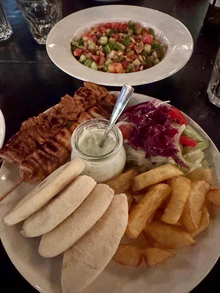 A hearty platter of grilled meat skewers, warm pita bread, thick-cut fries, a jar of tzatziki sauce, and a side salad, accompanied by a bowl of freshly chopped tomato and cucumber salad.
