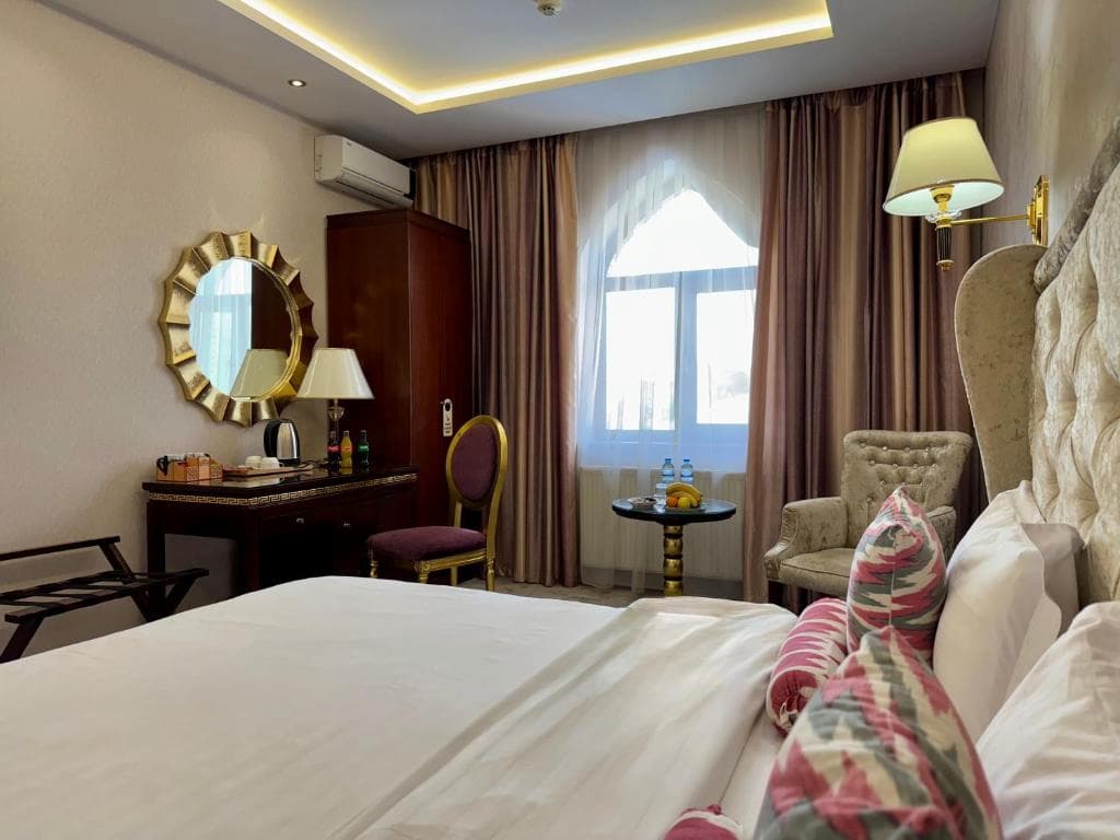 Classic hotel room at EmirHan Hotel in Samarkand featuring a double bed with patterned bolsters, a sunburst wall mirror, armchairs, and curtains framing an arched window.