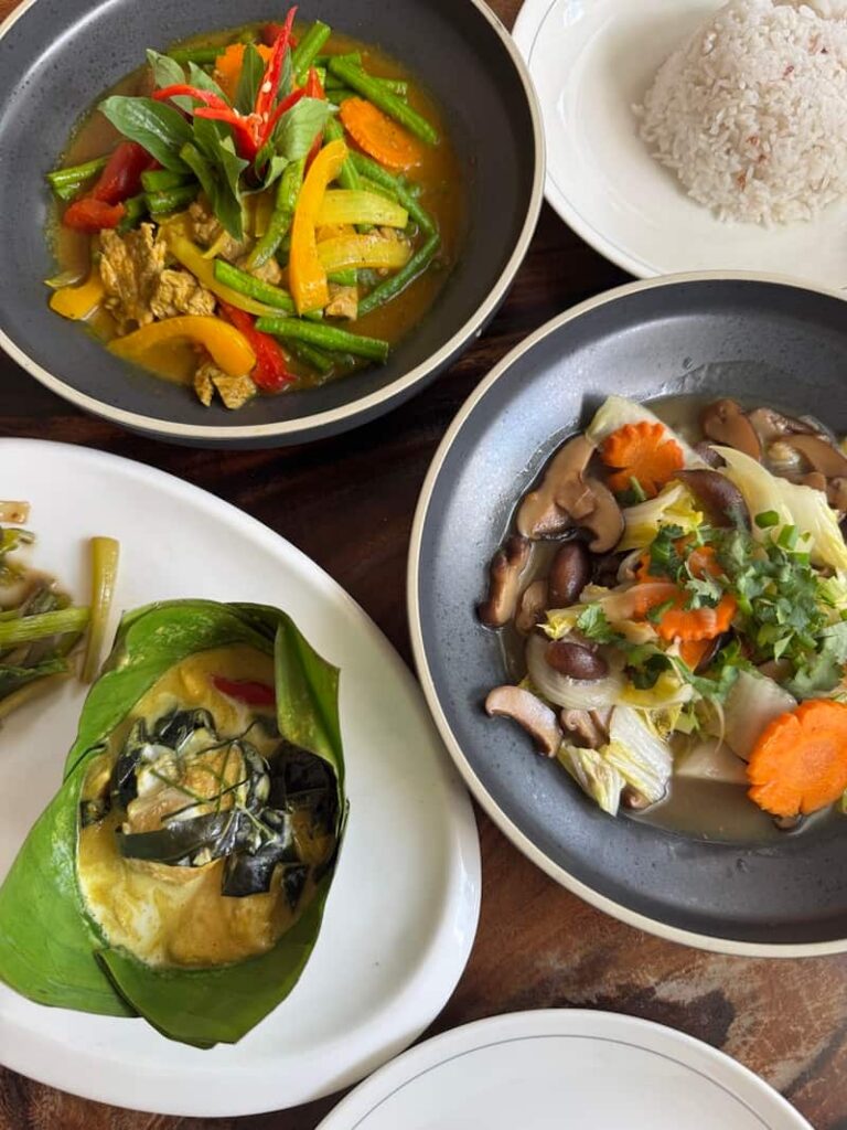 A spread of traditional Cambodian dishes, including fish amok in a banana leaf cup and vegetable stir-fries served with rice.