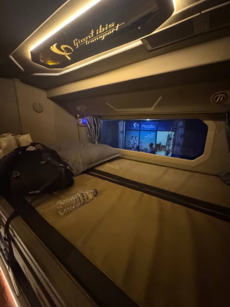 Interior view of a cozy double sleeper bunk with a pillow, backpack, and water bottle on a Giant Ibis bus.