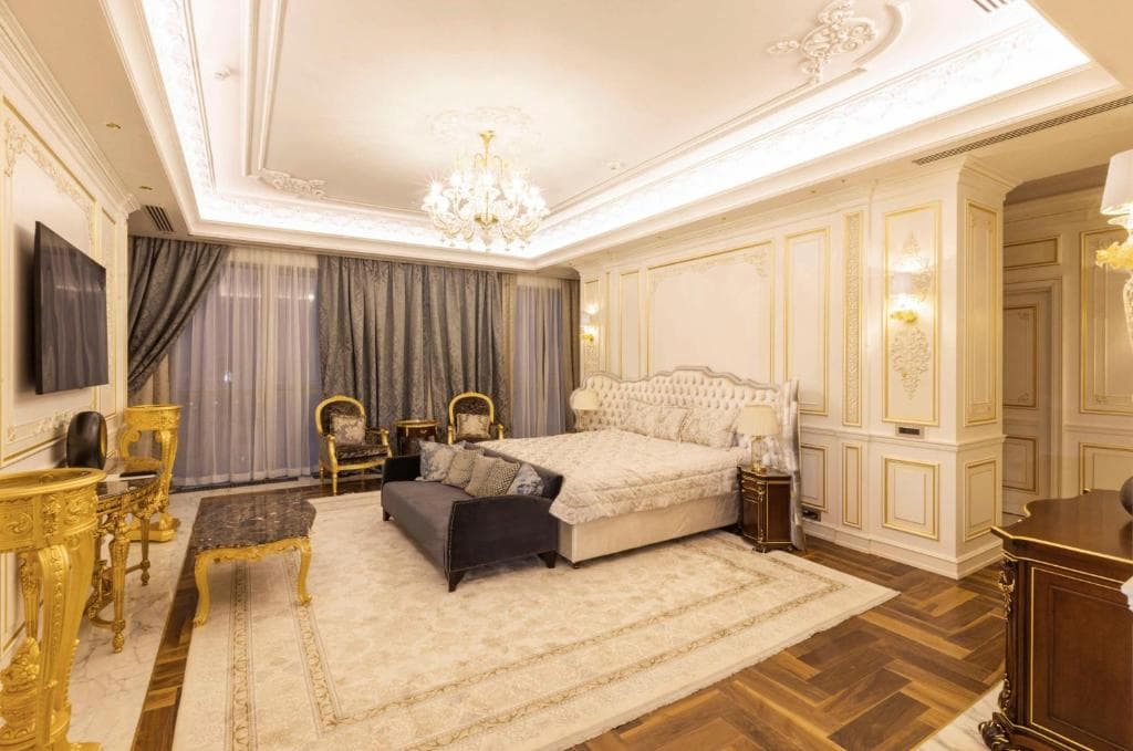 Luxurious hotel room at the Hilton in Samarkand featuring classic white and gold moldings, an ornate chandelier, a large bed with a sofa bench, and golden furniture accents.