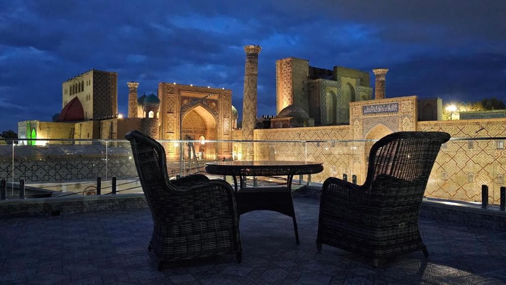 Rooftop terrace view of the illuminated Registan Square at night from Hotel Minor in Samarkand, featuring rattan chairs and a table.