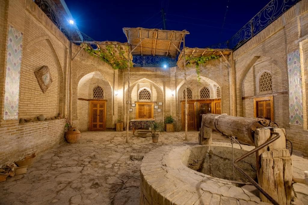 Traditional courtyard of the Khurjin Old Caravanserai Hotel in Bukhara at night, featuring historic brick archways, a stone well, and cobblestone ground.