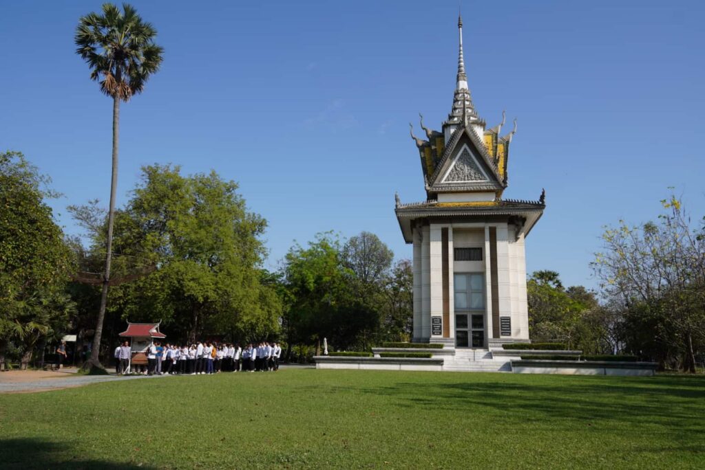 A wide-angle exterior view of the towering memorial stupa at the Choeung Ek Killing Fields, with visitors gathered nearby on a bright sunny day.