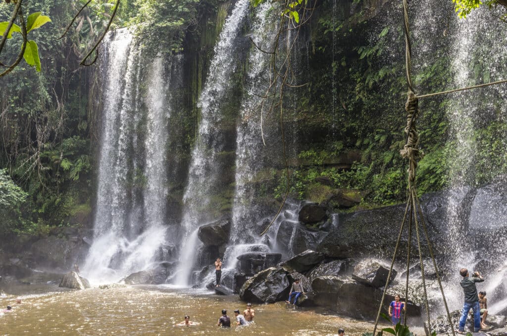People swimming and relaxing in the pool beneath the cascading waterfall at Phnom Kulen National Park near Siem Reap, Cambodia.