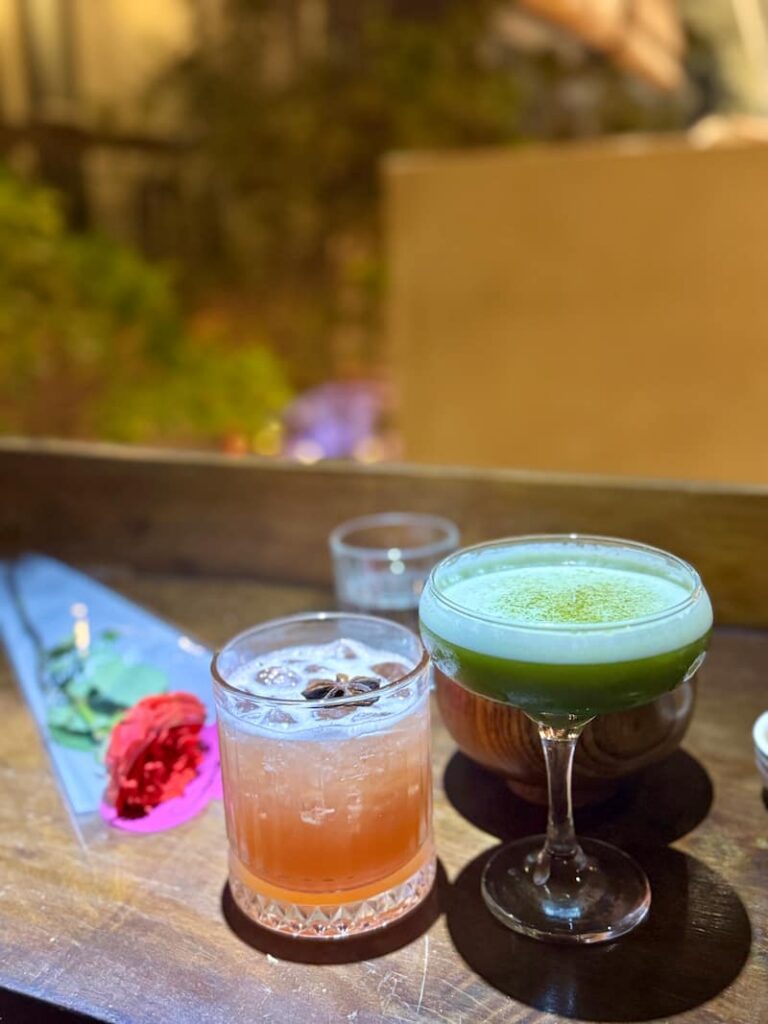 Two colorful cocktails—a green drink in a coupe glass and a garnished red cocktail with star anise next to a red rose—on a wooden bar table.