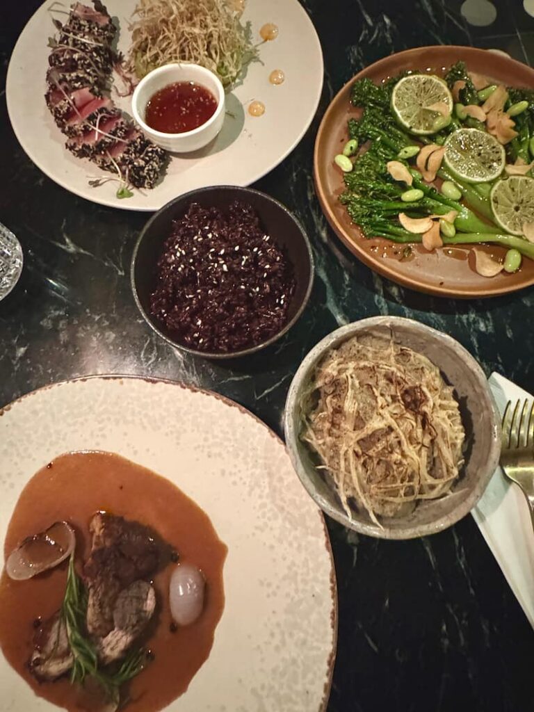 A spread of gourmet dishes, including sesame-crusted seared tuna, garlic broccolini, and black rice, served on a dark table at Le Langka restaurant in Phnom Penh.
