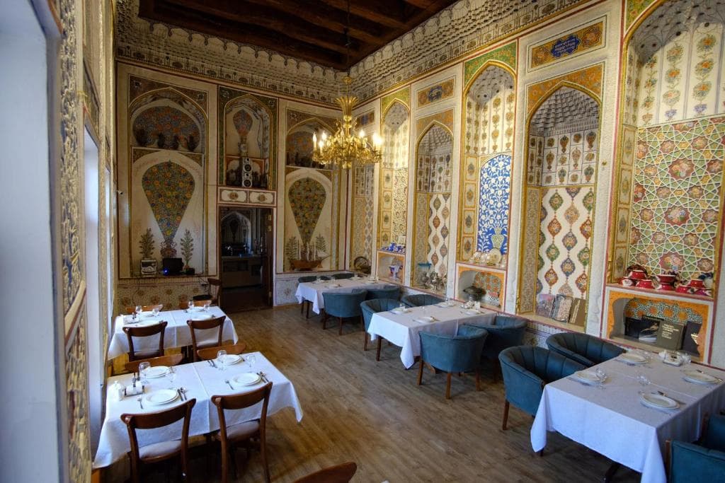Ornate dining room at Lyabi House Hotel in Bukhara featuring intricately painted traditional walls with mosaic niches, wooden floors, and white-clothed dining tables with a chandelier overhead.