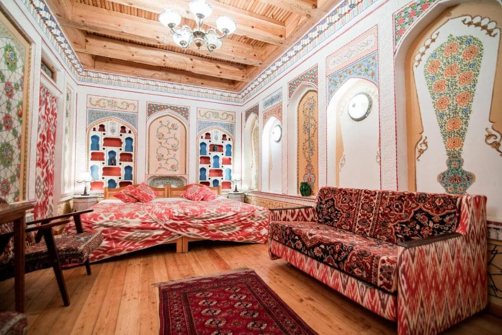 Traditional heritage room at Minzifa Boutique Hotel in Bukhara, featuring hand-painted walls with floral motifs, ornate niches, red ikat textiles, and a wood-beamed ceiling.