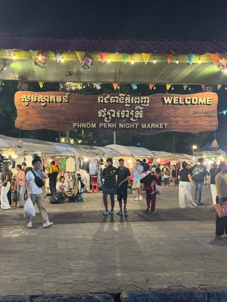 The illuminated wooden entrance sign reading 'Welcome Phnom Penh Night Market' above a bustling walkway filled with crowds and clothing stalls at night.