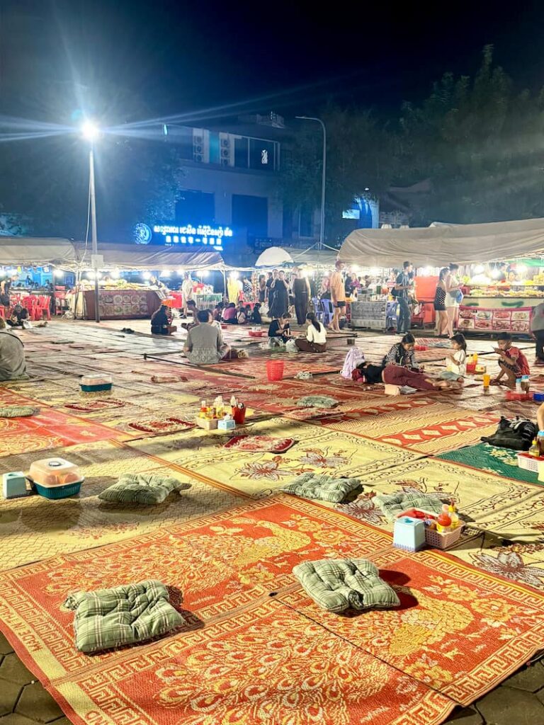 Colorful woven mats and floor cushions laid out on the ground for casual street food dining at the Phnom Penh Night Market, with food stalls and visitors in the background.