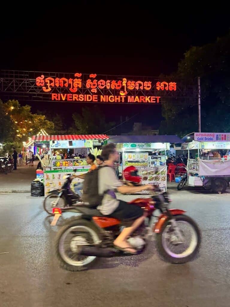A person riding a motorcycle past the illuminated entrance sign of the Riverside Night Market in Siem Reap at night, as shown in our guide with things to do in Siem Reap beyond Angkor Wat.