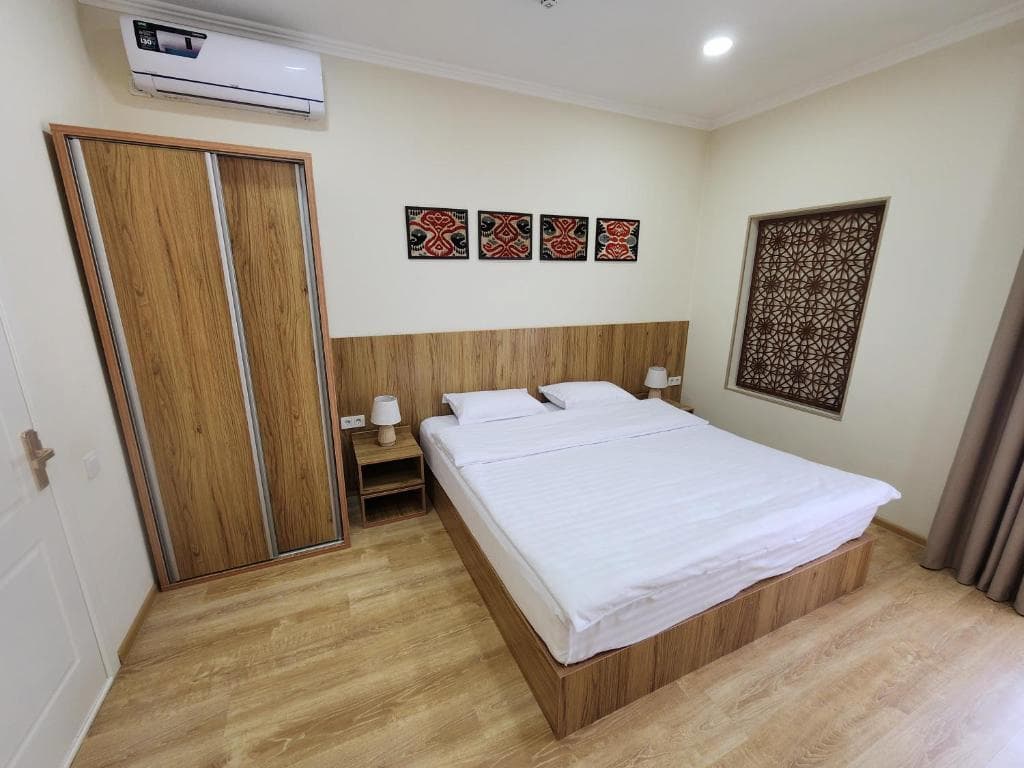Cozy hotel room at Oasis Boutique Hotel in Bukhara featuring a double bed, wooden furnishings, traditional framed textile art, and an ornate lattice window screen.