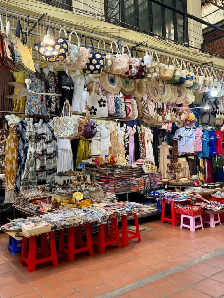 A vibrant market stall inside the Old Market in Siem Reap displaying hanging tote bags, straw purses, and clothing, as shown in our guide with things to do in Siem Reap beyond Angkor Wat.
