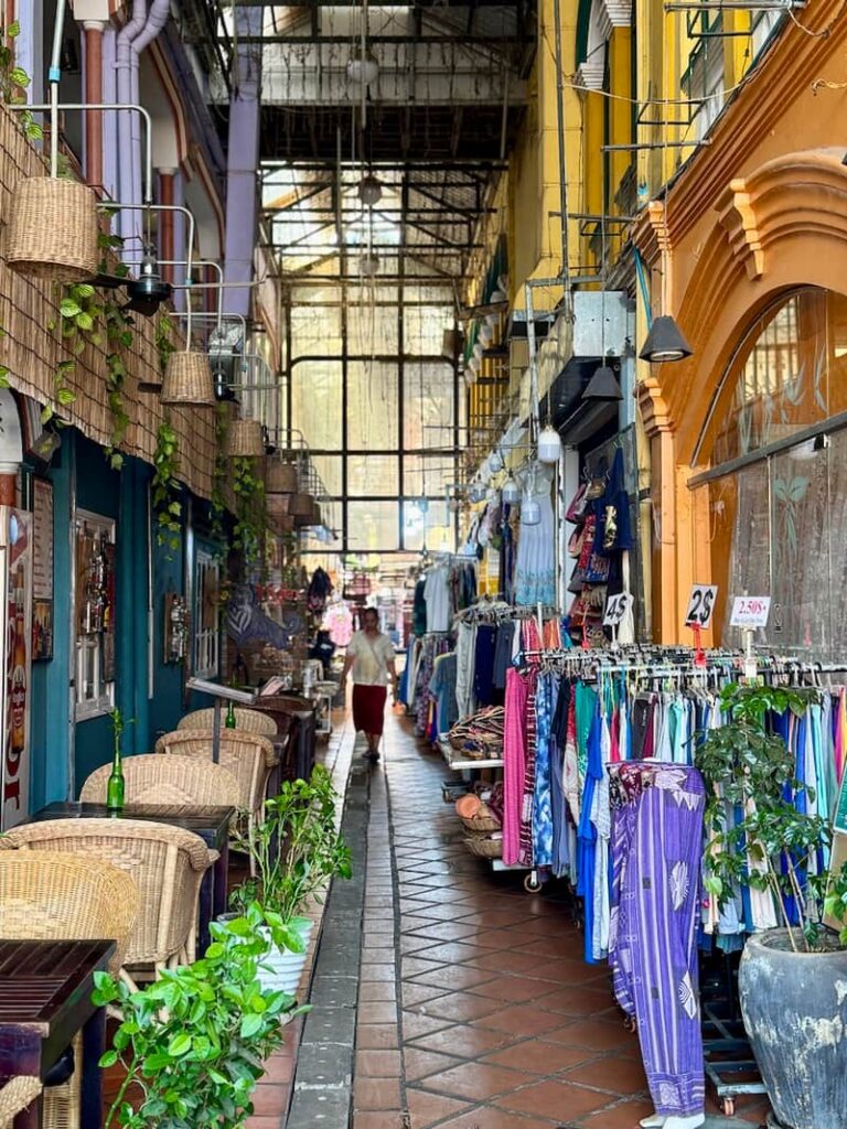 A narrow covered aisle of the Old Market in Siem Reap with clothing racks, outdoor cafe seating, and historic architecture.