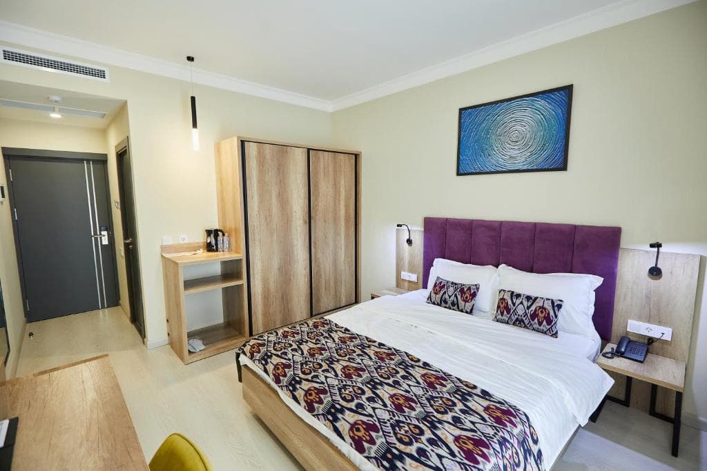 Comfortable hotel room at Old Tashkent Hotel and Spa featuring a double bed with a purple headboard, patterned traditional textiles, and wooden furnishings.