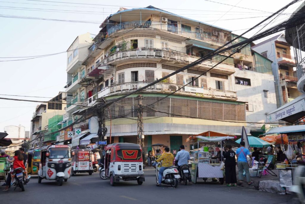Busy street scene in Phnom Penh with tuk-tuks, motorbikes, street food stalls, and an old multi-story building.