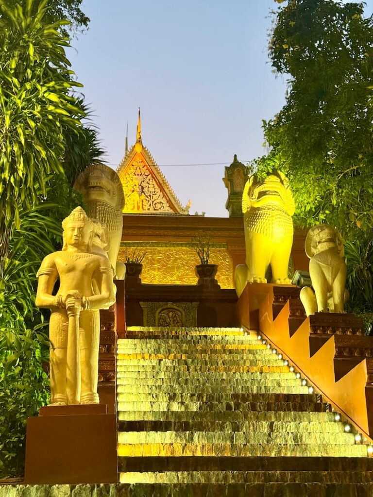 An illuminated stone staircase flanked by traditional guardian and lion statues leading up to a glowing temple at dusk in Phnom Penh.