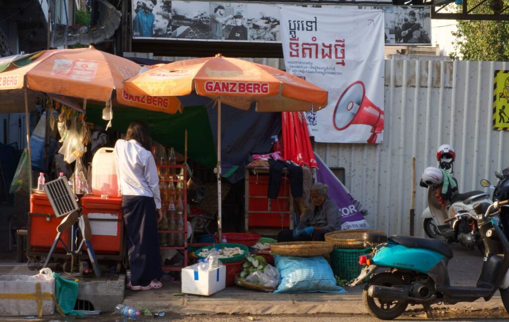A local street market stall shaded by orange Ganzberg umbrellas in Phnom Penh, featuring a vendor with baskets of produce, a customer browsing, and a teal scooter parked on the sidewalk.