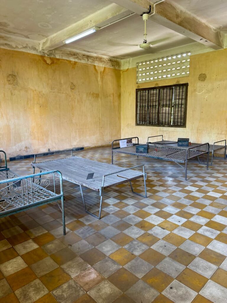 A sparse prison room at S-21 featuring metal bed frames on a checkerboard-tiled floor and stained yellow walls.