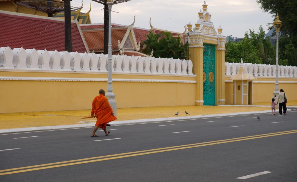 A Buddhist monk in vibrant orange robes walking past the Royal Palace wall, highlighting the culture that answers: Is Phnom Penh worth visiting?