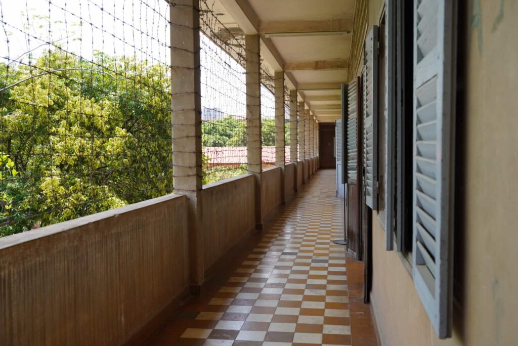 A long, tiled corridor at the Tuol Sleng Genocide Museum enclosed with security netting along the open pillars.