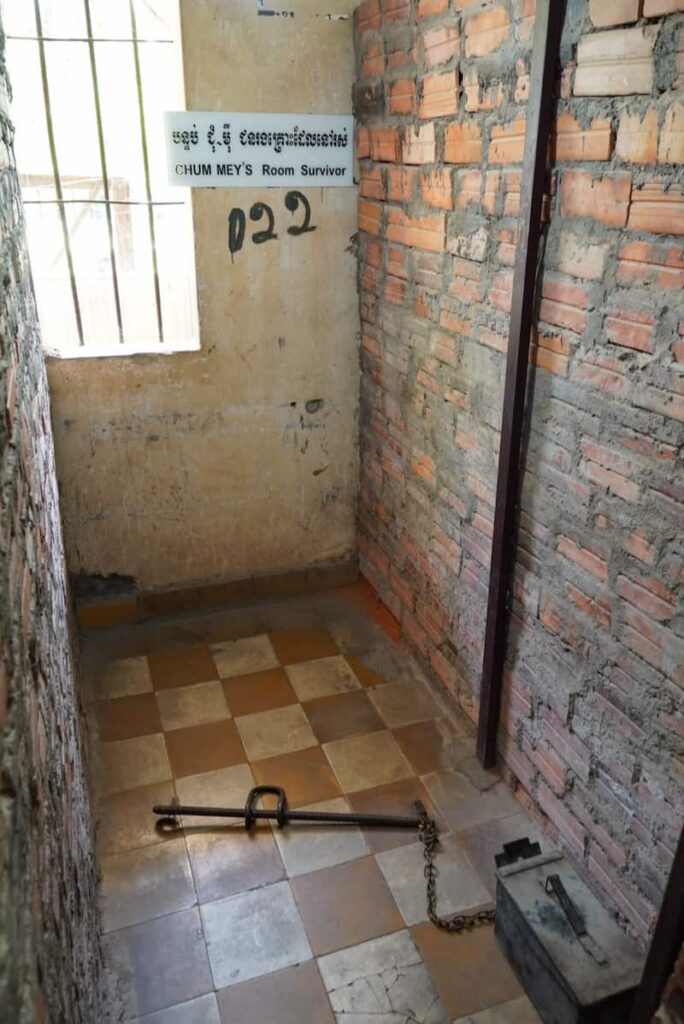 A small, stark prison cell at Tuol Sleng featuring a shackle on the checkerboard floor and a sign for survivor Chum Mey.