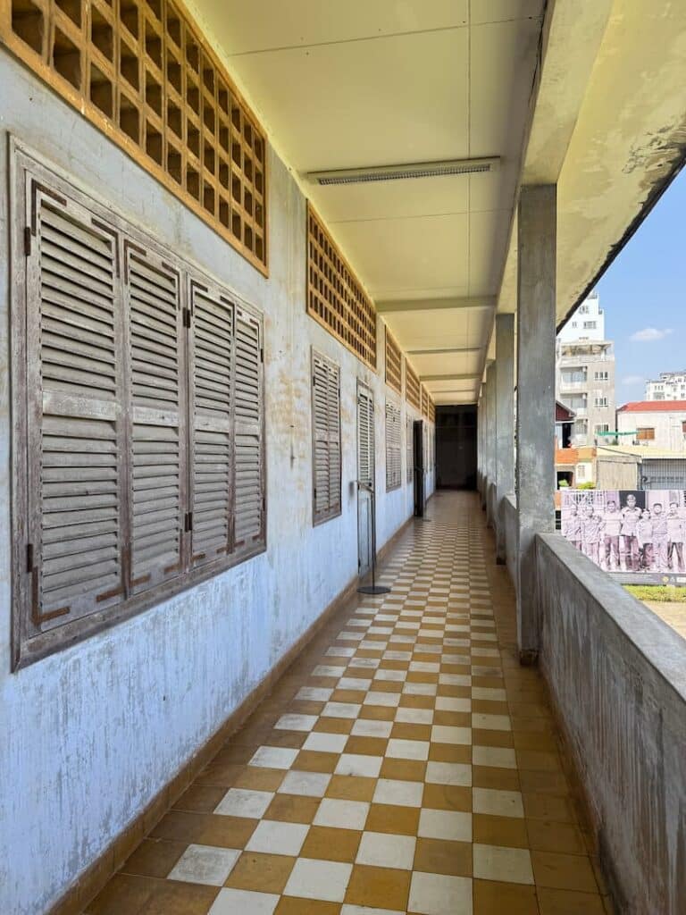 An exterior hallway at Tuol Sleng with a checkerboard-tiled floor, open concrete pillars, and wooden shuttered windows as shown in our Phnom Penh Killing Fields and S21 respectful guide.