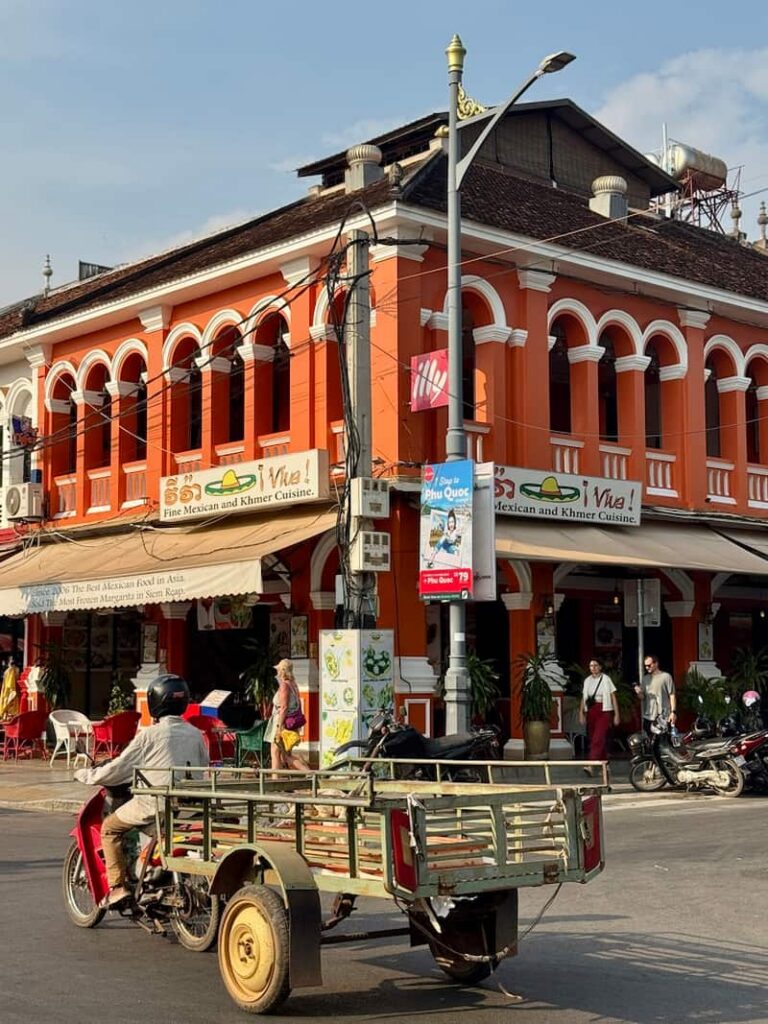 A red motorized tuk-tuk driving past a bright orange colonial building and restaurant in Siem Reap, Cambodia.