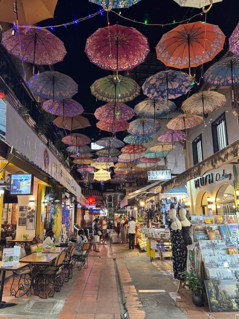 A canopy of colorful, patterned umbrellas and string lights hanging overhead along a bustling pedestrian alley in the central market of Siem Reap at night.