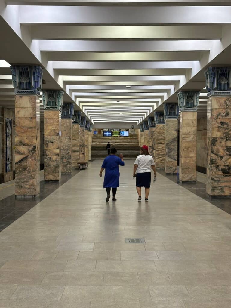 Two people walking down the platform of an ornate Tashkent metro station with marble pillars and a geometric ceiling.